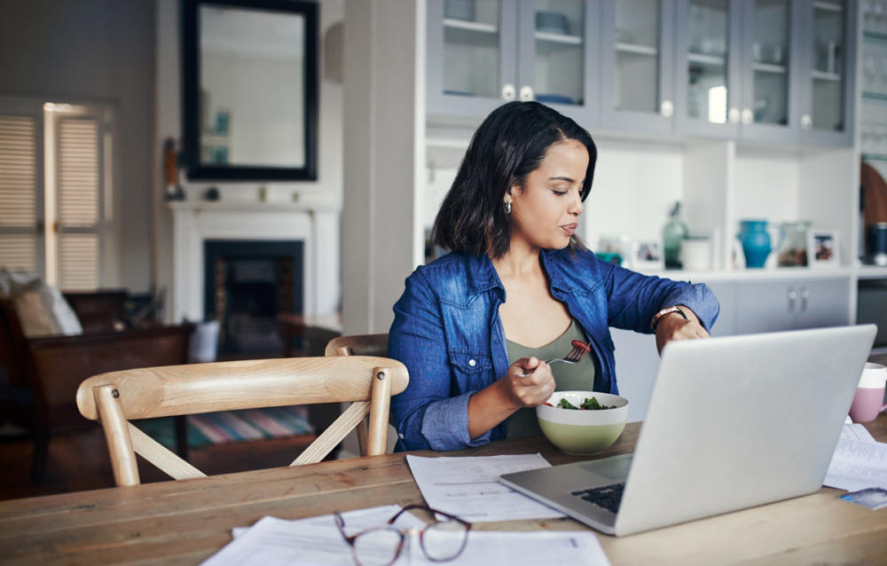 Woman eating at kitchen table while working on computer