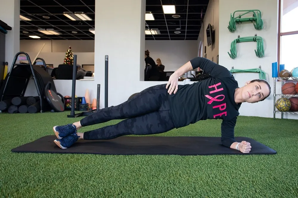 Female performing a side plank on a mat in a gym.